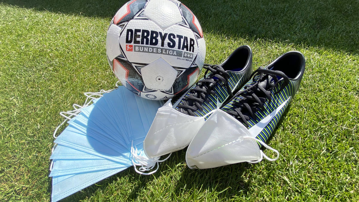 Ein Bundesliga-Spielball, ein Paar Fußballschuhe und aufgefächerte medizinische Masken liegen auf dem Rasen
