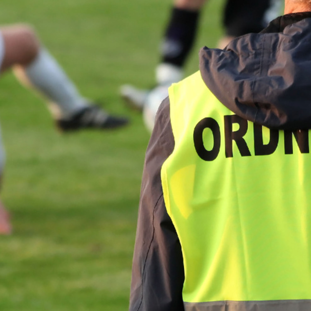 Ein Ordner in gelber Warnweste am Spielfeldrand, im Hintergrund unscharf zwei Spieler