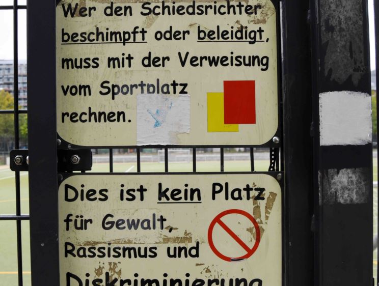 Zwei Schilder an einem Sportplatzzaun: „Wer den Schiedsrichter beschimpft oder beleidigt, muss mit der Verweisung vom Sportplatz rechnen“ mit gelber und roter Karte, darunter „Dies ist kein Platz für Gewalt, Rassismus und Diskriminierung“